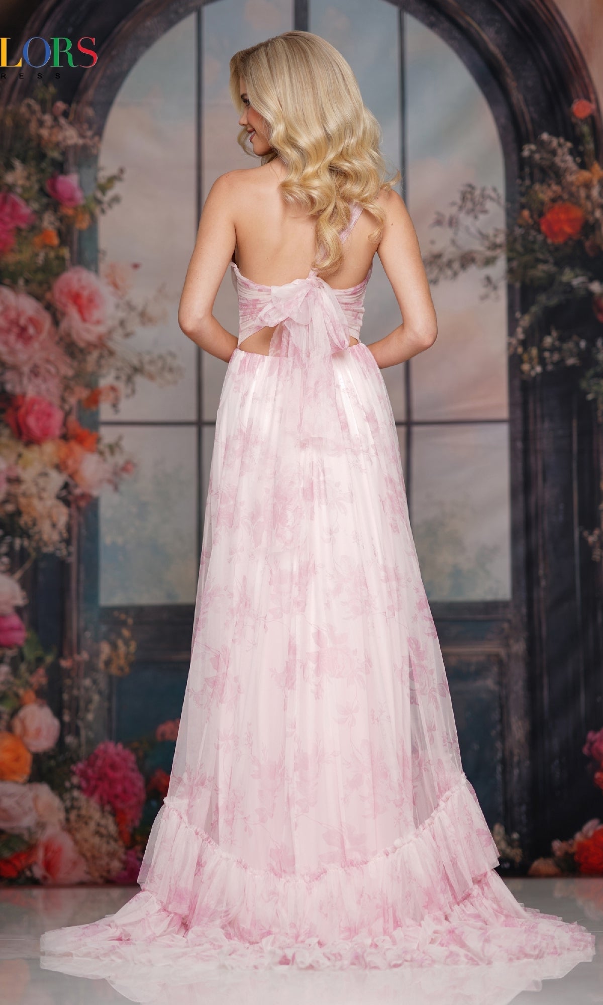 A woman in the Colors Dress 3814, a long light pink chiffon prom dress with a back bow, stands facing away before a floral arched window backdrop.